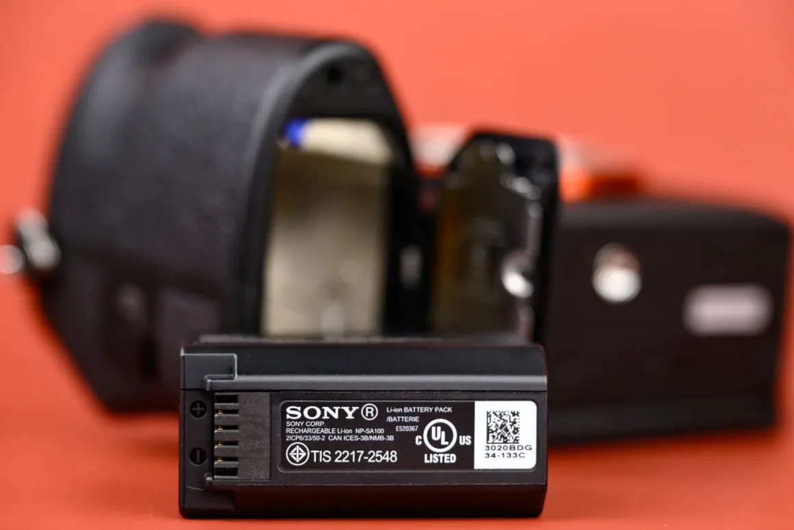 Turuncu arka plan üzerinde, bulanık bir dijital kameranın açık pil yuvası önünde duran Sony kamera pili.