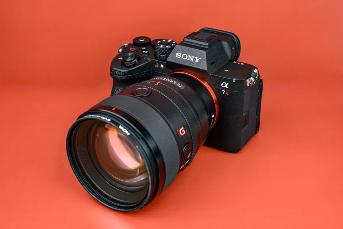 Turuncu arka plan üzerinde sergilenen büyük Sony G Master lens takılmış siyah Sony α7R aynasız kamera.