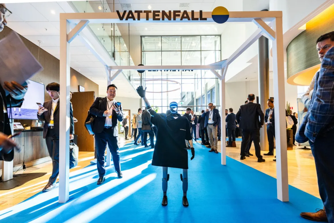 İş kıyafetli insanların bulunduğu kalabalık bir kapalı etkinlikte, “Vattenfall” yazılı beyaz bir çerçevenin altında duran insansı bir robot. Bazı katılımcılar etkileşim halinde, bazıları telefonlarını kullanıyor.