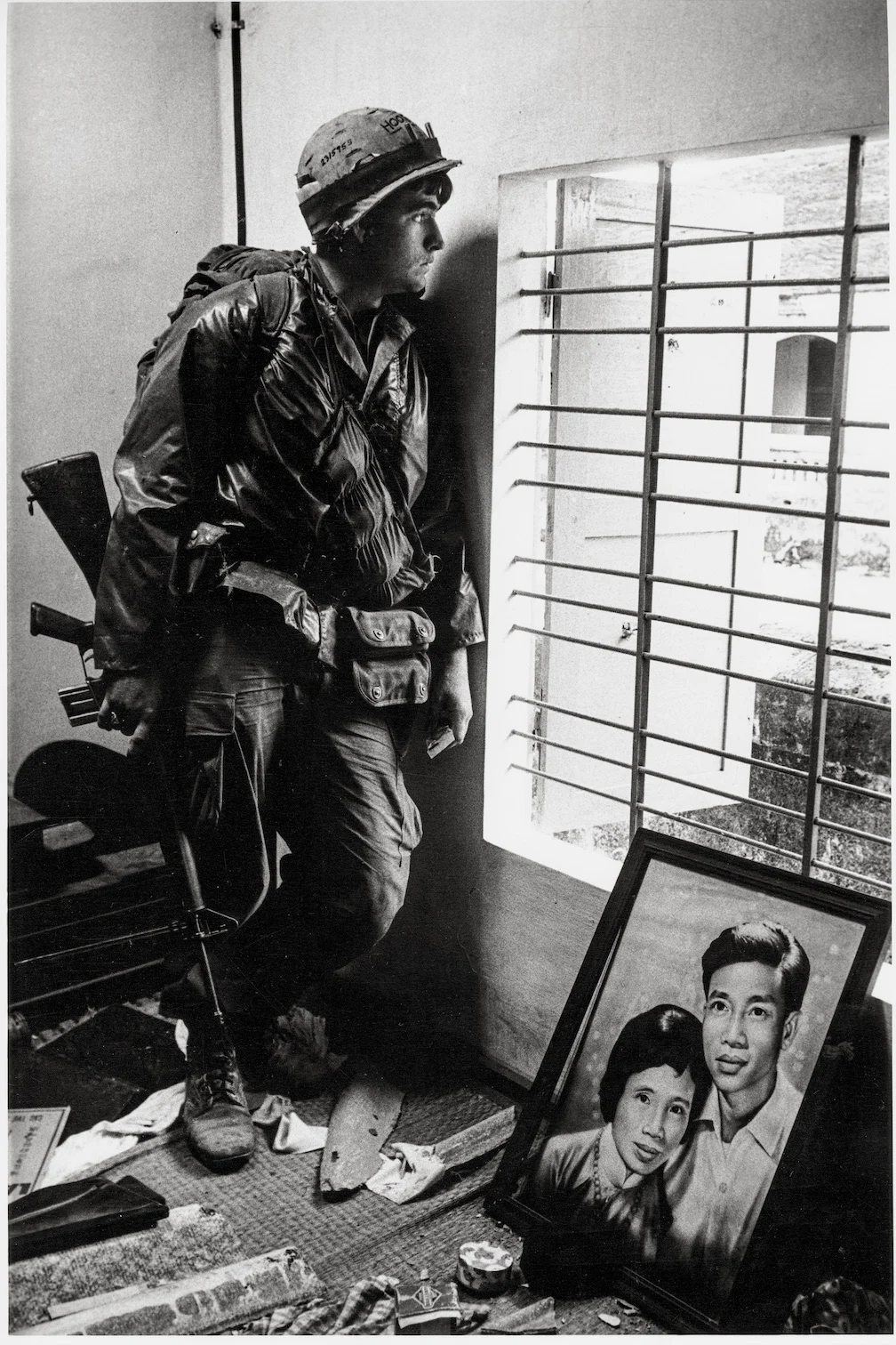 Don McCullin’in Son Fotoğraf Kitabı Vietnam’a Yaptığı 16 Yolculuğu Bir Araya Getiriyor 2 Üniformalı ve kasklı bir asker, tüfeğini tutarak parmaklıklı bir pencerenin yanında duruyor. Yanında yerde bir kadın ve erkeğin çerçeveli portresi ile etrafa dağılmış eşyalar bulunuyor.