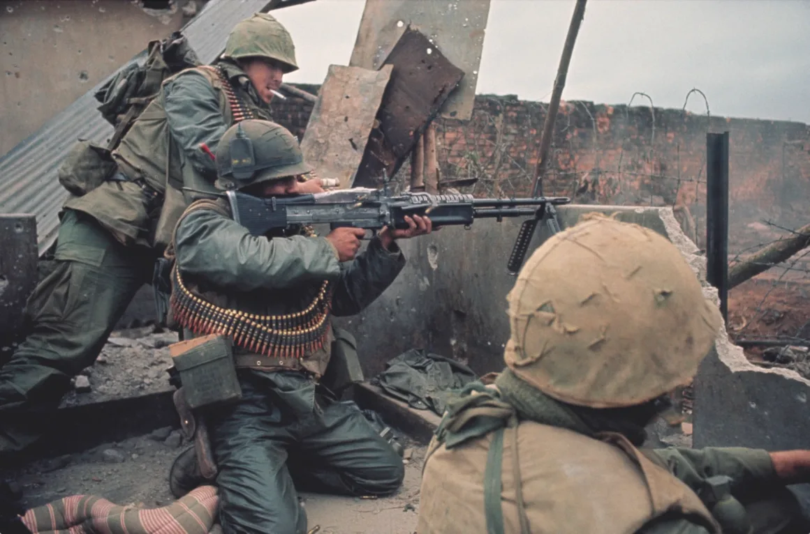 Don McCullin’in Son Fotoğraf Kitabı Vietnam’a Yaptığı 16 Yolculuğu Bir Araya Getiriyor 3 Savaş teçhizatı taşıyan üç asker molozların arkasına sığınmış durumda. Biri makineli tüfeğini nişan alırken, diğeri ayakta bekliyor ve üçüncü asker çömelmiş halde bulunuyor. Etraflarında yıkılmış duvarlar ve savaşın izleri görülüyor.