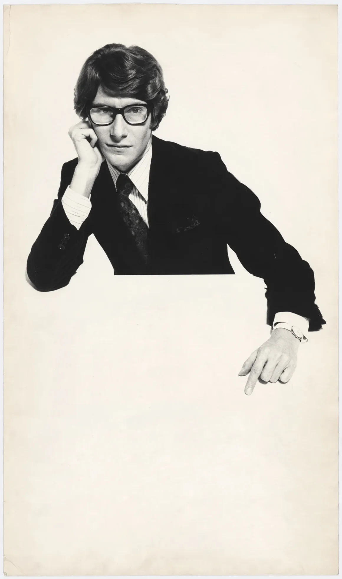 Yves Saint Laurent ve Fotoğraf: Bir Moda Efsanesini Şekillendiren 40 Yıl 4 Takım elbise ve gözlük takan bir adam, sade beyaz bir arka plan önünde başını bir eline yaslamış halde diğer eliyle aşağıyı işaret ediyor.