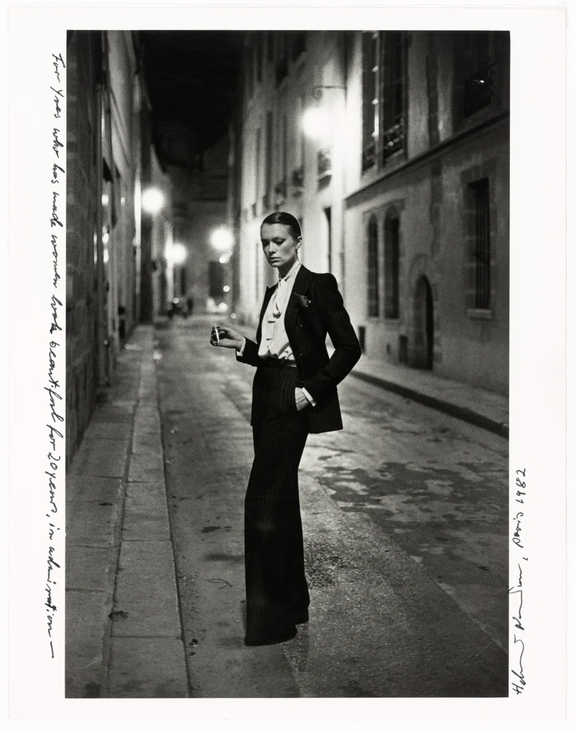 Yves Saint Laurent ve Fotoğraf: Bir Moda Efsanesini Şekillendiren 40 Yıl 5 Çizgili bir takım elbise ve papyon giyen kişi, gece vakti loş ışıklarla aydınlanan boş bir şehir sokağının ortasında elinde sigarayla kendinden emin bir şekilde duruyor. Fotoğrafın iki kenarında el yazısıyla notlar bulunuyor.
