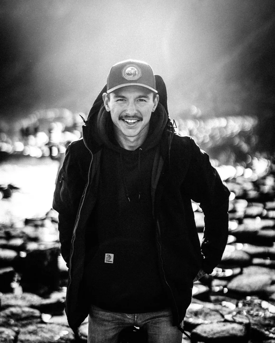 Jake Guzman: Otherworldly America 2 Şapka ve ceket giyen bir adam, elleri ceplerinde kameraya gülümserken açık havada duruyor. Arka plandaki kayalık ve güneş ışığı alan ortam dramatik bir ışık etkisi yaratıyor. Fotoğraf siyah beyaz.