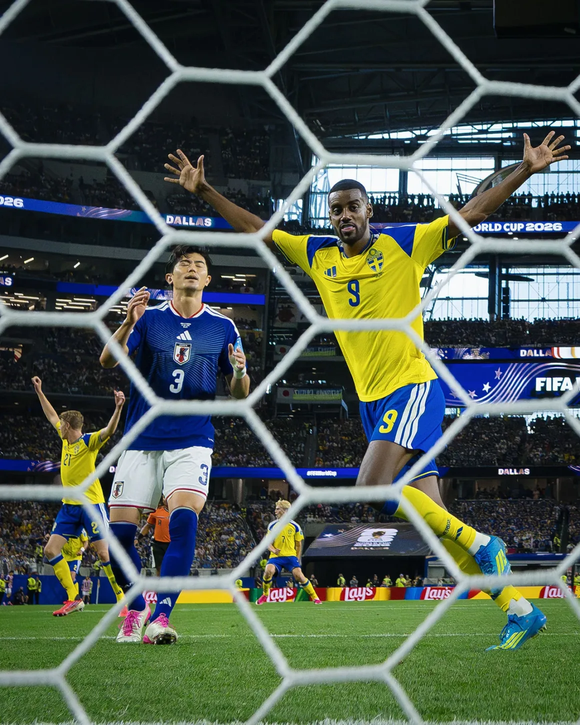 İki Spor Fotoğrafçısı Dünya Kupası’nı Görüntülemenin Nasıl Bir Deneyim Olduğunu Anlatıyor 24 Sarı İsveç forması giyen bir futbolcu kollarını kaldırarak gol sevincini yaşarken, yakınındaki Japon savunma oyuncusu tepki gösteriyor. Sahne, dolu bir stadyumda kale ağlarının ardından görüntüleniyor.