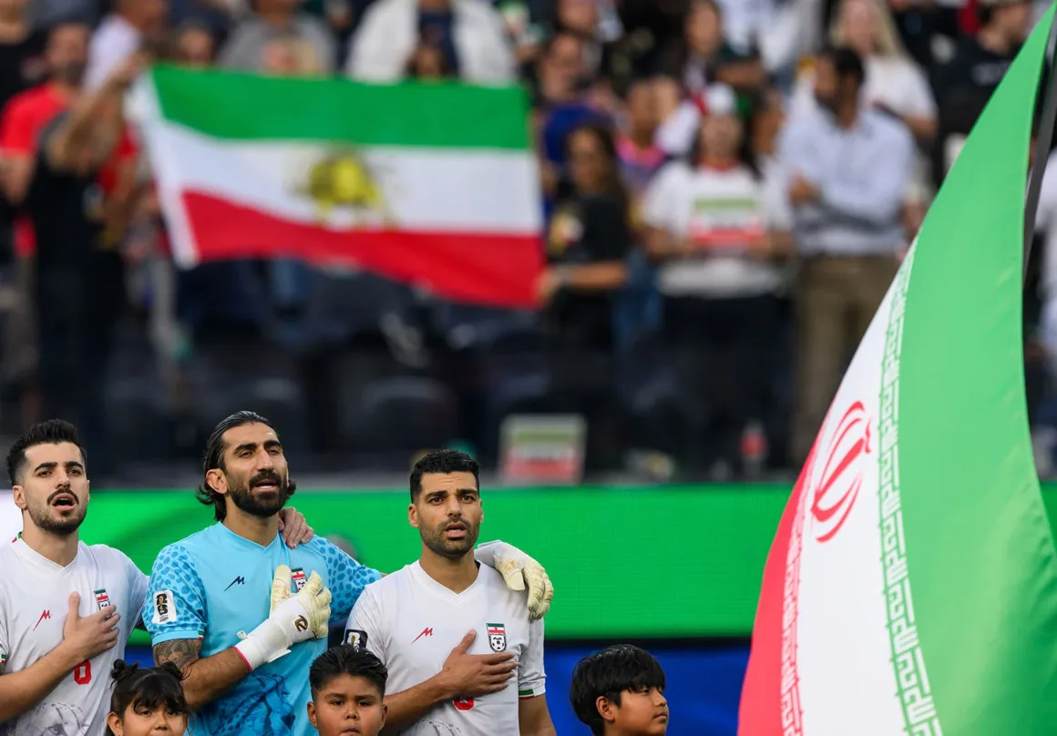 İki Spor Fotoğrafçısı Dünya Kupası’nı Görüntülemenin Nasıl Bir Deneyim Olduğunu Anlatıyor 5 Üç İranlı futbolcu milli marş sırasında ellerini göğüslerine koymuş halde duruyor. Ön planda büyük bir İran bayrağı, arka planda ise tribünlerde başka bir İran bayrağı taşıyan taraftarlar görülüyor.