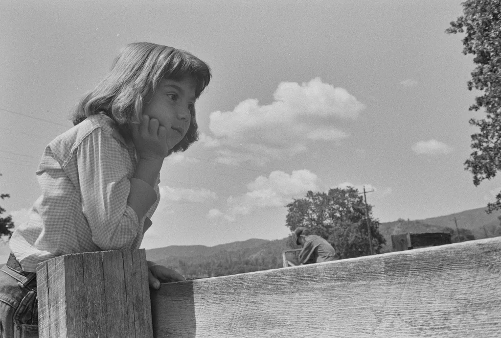Dorothea Lange'ın Sular Altında Kalan Kasabayı Belgeleyen Fotoğrafları Gün Yüzüne Çıktı 4 Ahşap bir çite yaslanan genç bir kız, çenesini eline dayamış şekilde düşünceli biçimde uzaklara bakıyor. Arka planda ağaçlar, bulutlar ve dağlar görülüyor.