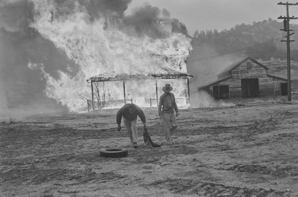 Dorothea Lange'ın Sular Altında Kalan Kasabayı Belgeleyen Fotoğrafları Gün Yüzüne Çıktı 8 İki adam alevler içindeki bir evden uzaklaşırken gökyüzüne yoğun duman yükseliyor. Yakındaki ahşap bir bina da yangının yanında duruyor. Çorak arazi ve dramatik atmosfer dikkat çekiyor.
