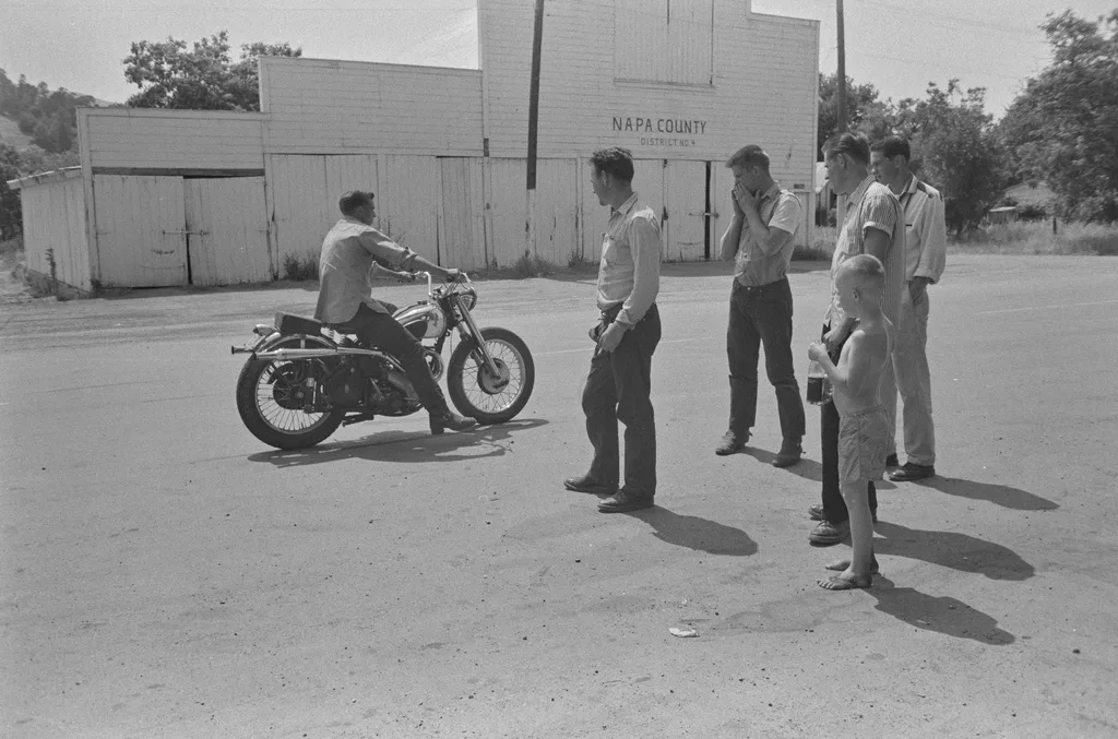 Dorothea Lange'ın Sular Altında Kalan Kasabayı Belgeleyen Fotoğrafları Gün Yüzüne Çıktı 5 Bir adam motosiklet sürerken dört erkek ve üstü çıplak bir çocuk onu izliyor. Arka planda "Napa County Districts" yazılı beyaz ahşap binalar bulunuyor. Sahne 20. yüzyılın ortalarına ait görünüyor.
