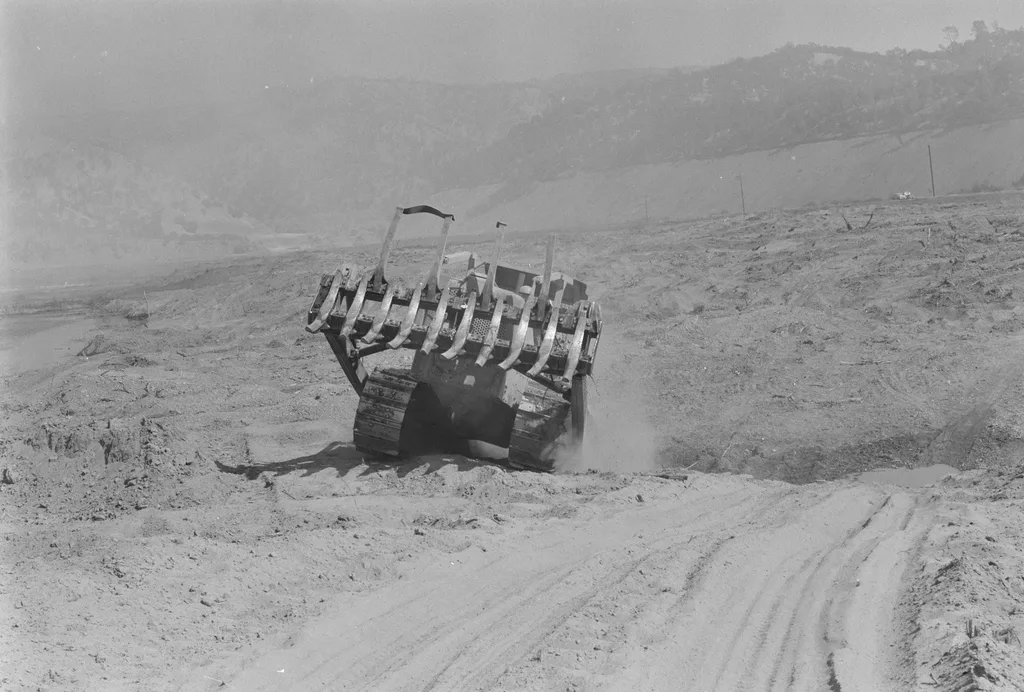 Dorothea Lange'ın Sular Altında Kalan Kasabayı Belgeleyen Fotoğrafları Gün Yüzüne Çıktı 7 Bir buldozer tozlu ve çıplak bir arazide toprağı kazıyor. Ön planda palet izleri ve toprak sırtları, arka planda ise tepeler ve puslu bir gökyüzü yer alıyor.