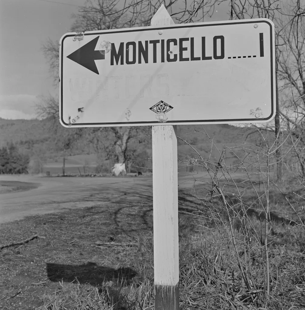 Dorothea Lange'ın Sular Altında Kalan Kasabayı Belgeleyen Fotoğrafları Gün Yüzüne Çıktı 3 Kırsal bir yolun kenarında sola yönü gösteren ve "Monticello ..... 1" yazılı bir yol tabelası. Arka planda çimenler, yapraksız ağaçlar ve tepeler yer alıyor.