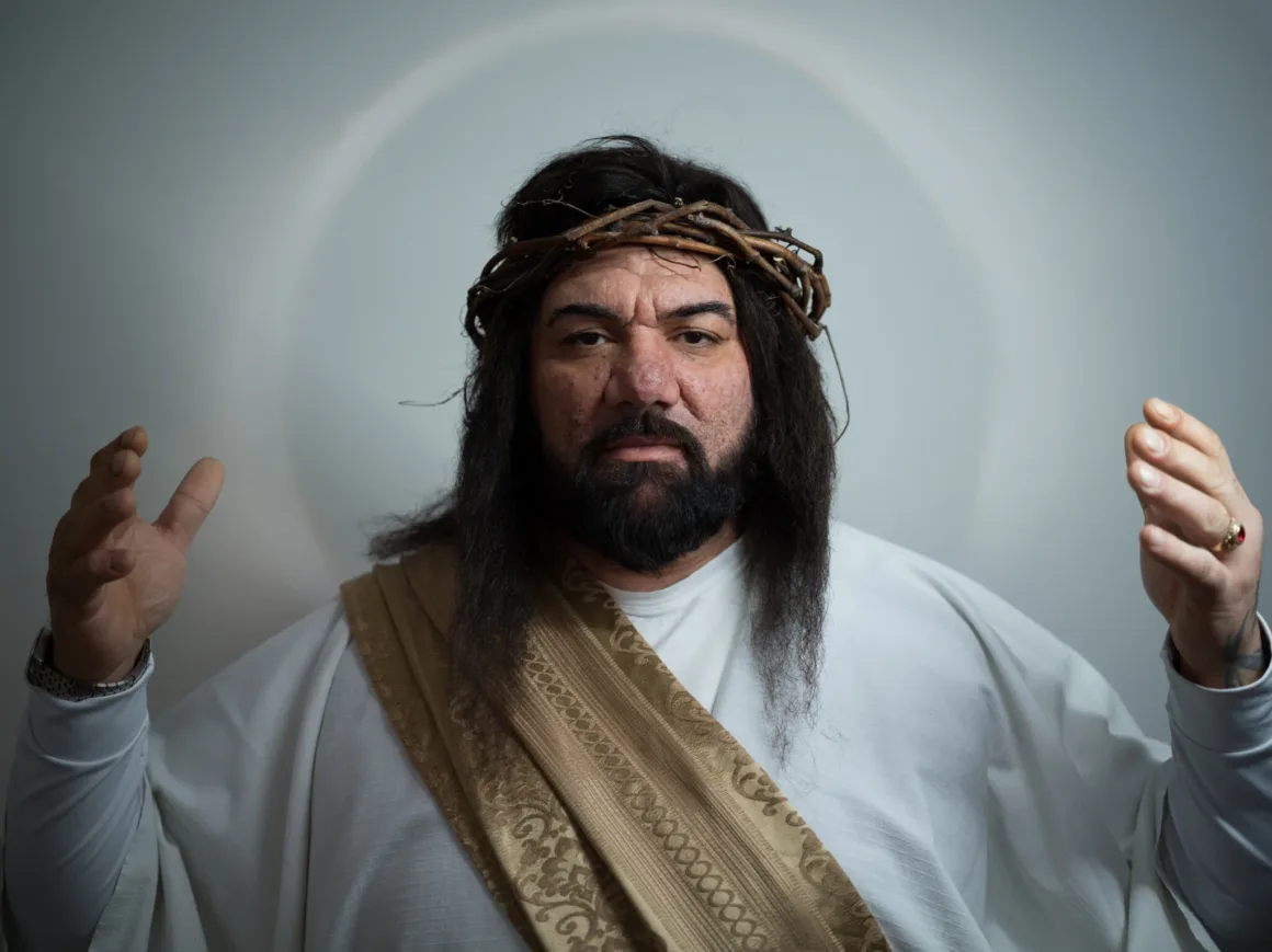 İncil dönemini andıran giysiler giyen ve dikenli taç takan bir kişi, başının arkasındaki hale benzeri daireyle birlikte iki elini havaya kaldırıyor. Görünüm, İsa'nın geleneksel tasvirlerini çağrıştırıyor.