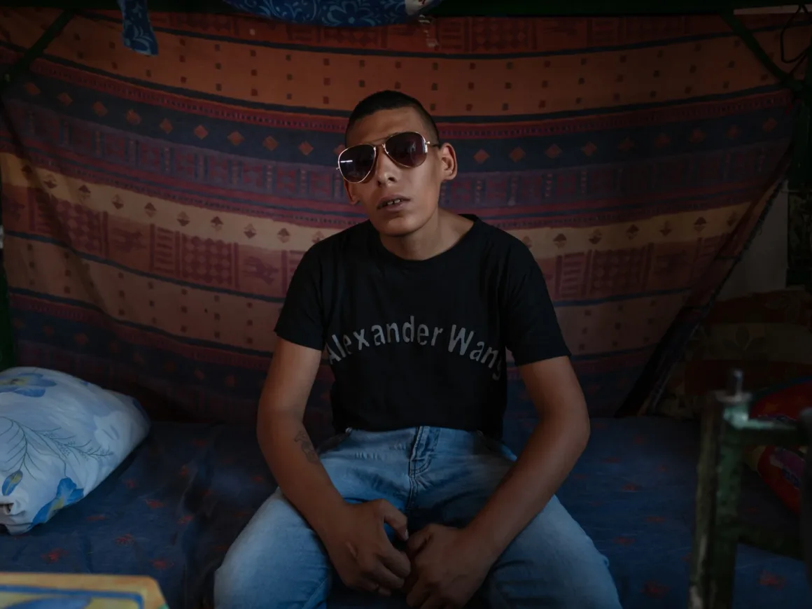 Güneş gözlüğü ve siyah Alexander Wang tişörtü giyen genç bir mahkûm, arkasında desenli bir battaniyenin asılı olduğu loş bir odada yatağın üzerinde oturuyor. Ellerini dizlerinin üzerine koymuş durumda.