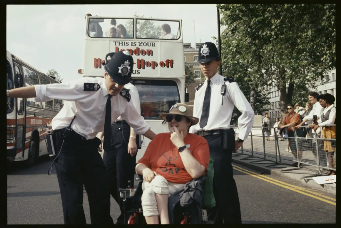Two Metropolitan Police officers in shirtsleeves direct a wheelchair user in a red t-shirt and sun hat away from a road blocked by buses during a Whitehall disability rights protest.