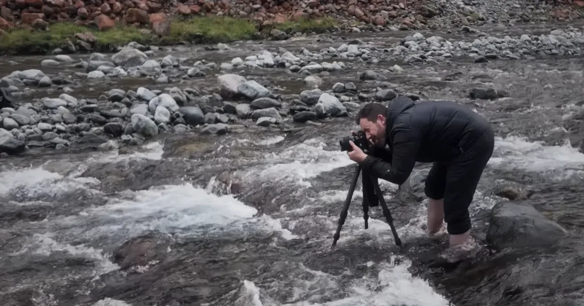 Michael Shainblum, Yeni Zelanda'nın Olağanüstü Güzelliğini Yeniden Yorumluyor 9 Siyah ceketli bir kişi, kayalık bir dere içinde çıplak ayakla durarak tripod üzerindeki kamerasını ayarlıyor. Su bacaklarının etrafından akarken kıyıda kayalar ve çimenler görülüyor.