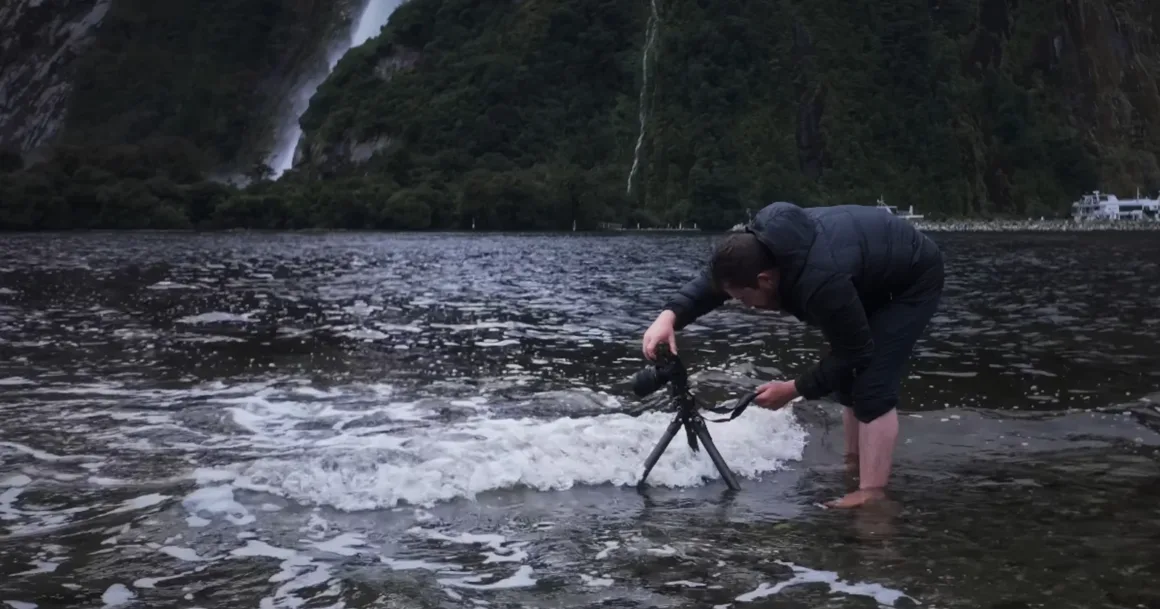 Michael Shainblum, Yeni Zelanda'nın Olağanüstü Güzelliğini Yeniden Yorumluyor 5 Çıplak ayakla sığ suda duran bir kişi, yaklaşan dalgalar arasında tripod üzerindeki kamerasını ayarlıyor. Arka planda ağaçlarla kaplı dağlar ve bir şelale yer alırken, kapalı gökyüzü sahneye kasvetli ve doğal bir atmosfer katıyor.