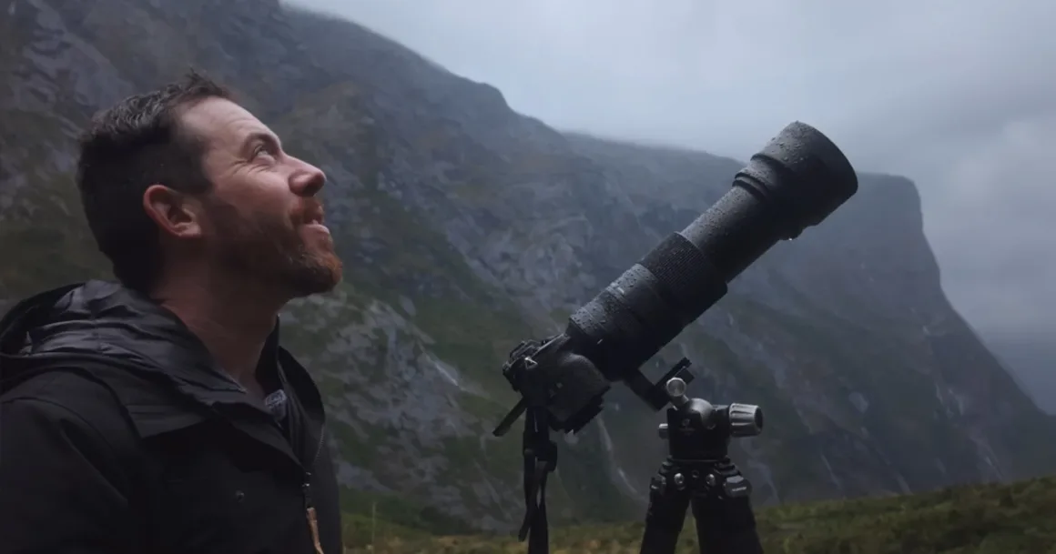 Michael Shainblum, Yeni Zelanda'nın Olağanüstü Güzelliğini Yeniden Yorumluyor 18 Sakallı bir adam, tripod üzerindeki telefoto objektifli büyük bir kameranın yanında açık havada duruyor ve arka plandaki sisli dağlara bakıyor.
