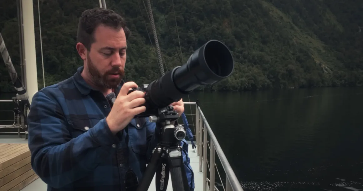 Michael Shainblum, Yeni Zelanda'nın Olağanüstü Güzelliğini Yeniden Yorumluyor 22 Mavi kareli gömlek giyen bir adam, ormanlarla kaplı tepeler ve sakin suyun önünde, tekne güvertesinde tripod üzerindeki telefoto objektifli büyük bir kamerayı ayarlıyor.