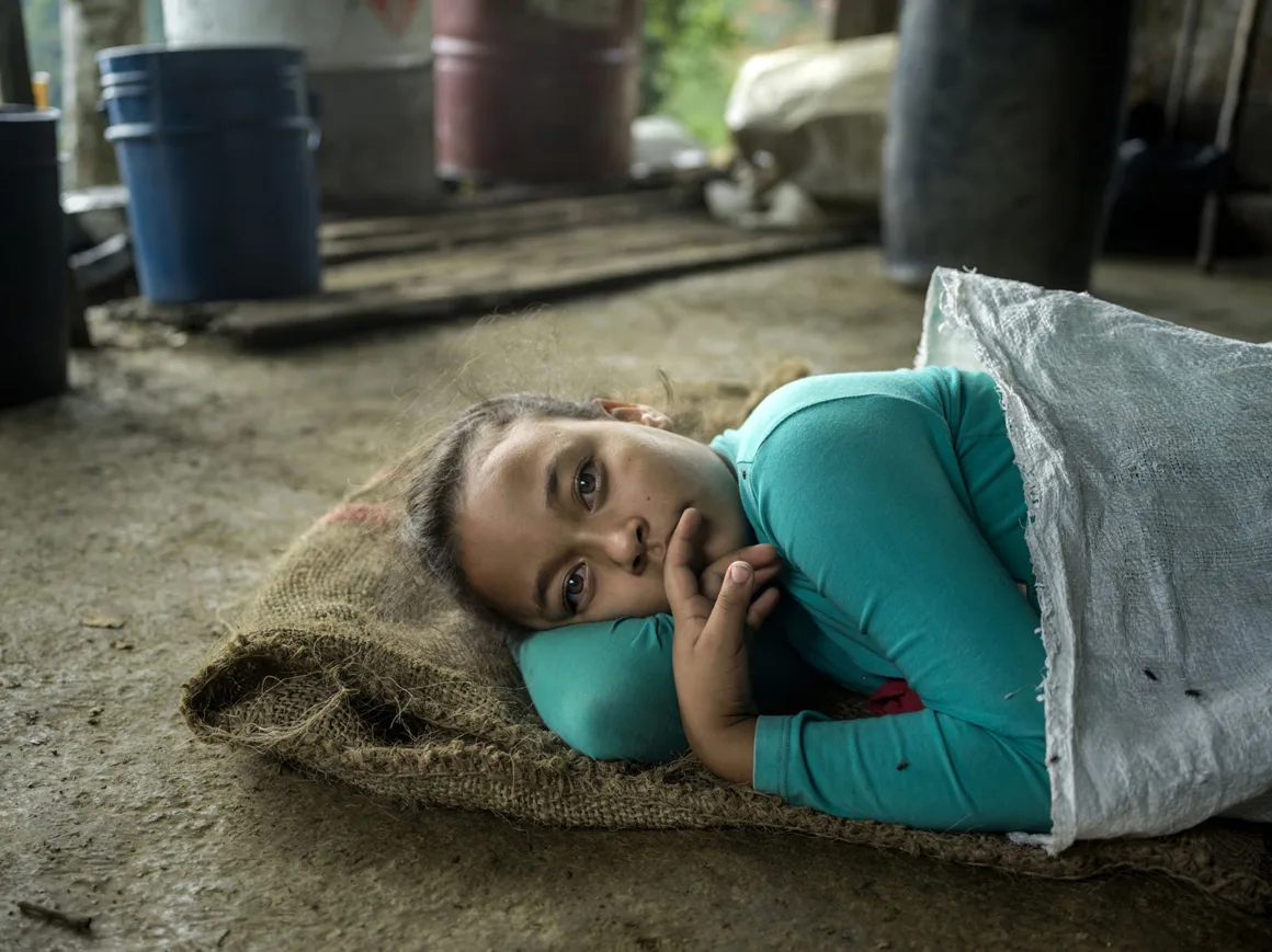 Turkuaz renkli tişört giyen genç bir kız, yerdeki çuvalın üzerinde beyaz bir örtüye kısmen sarılmış halde uzanıyor ve düşünceli bir ifadeyle kameraya bakıyor. Arka planda plastik variller ve ilkel bir laboratuvar ortamı görülüyor.