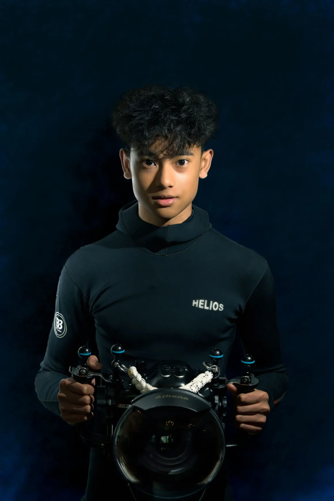 14 Yaşında Hasselblad Masters Kazandı: Su Altı Fotoğrafçısı Pari ile Tanışın 6 Koyu renk dalış kıyafeti giyen, kıvırcık saçlı genç bir fotoğrafçı, koyu mavi arka plan önünde profesyonel bir su altı kamerasını tutarak objektife bakıyor.