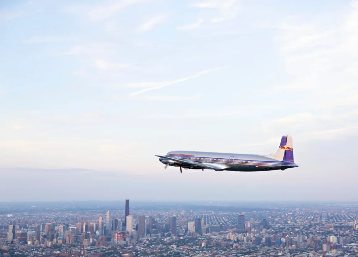 Red Bull, 68 Yıllık DC-6B'nin Tarihi Dönüşünü Vintage Bolex Kameralarla Belgeledi 4 Mavi gökyüzü ve hafif bulutlar altında geniş bir şehir silüetinin üzerinde uçan vintage pervaneli bir uçak.