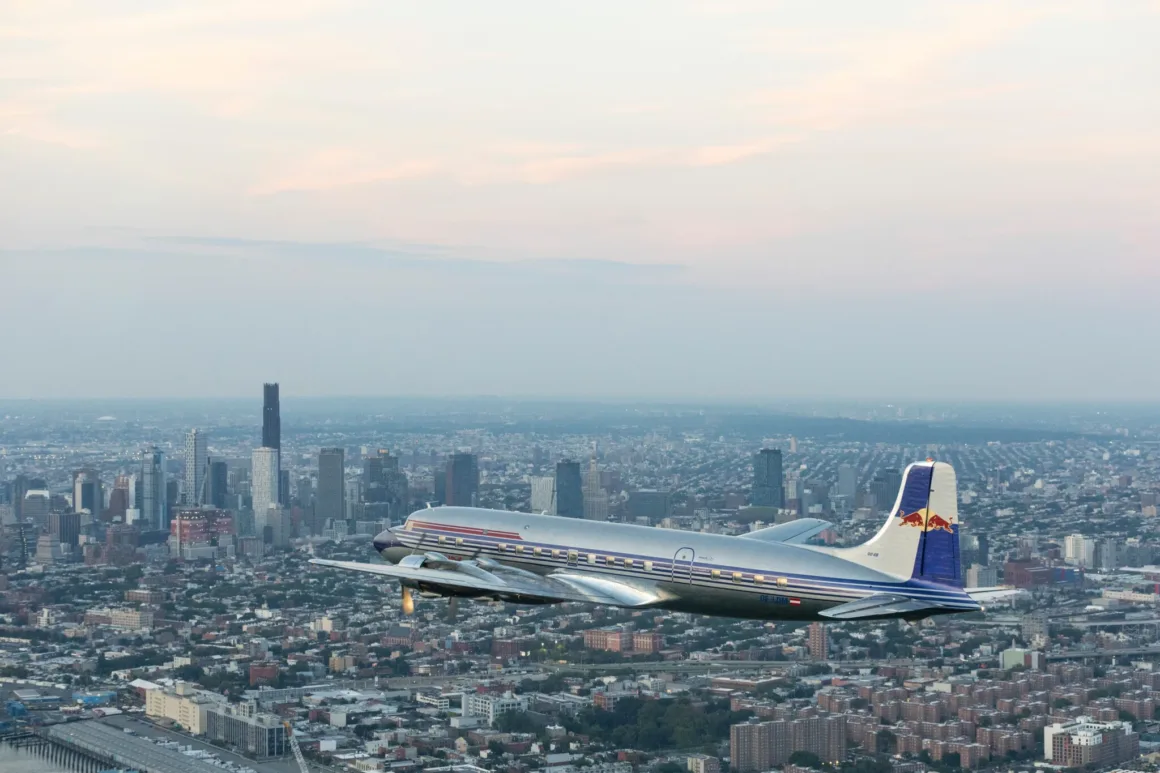 Red Bull, 68 Yıllık DC-6B'nin Tarihi Dönüşünü Vintage Bolex Kameralarla Belgeledi 3 Kırmızı ve mavi detaylara sahip gümüş renkli vintage bir uçak, bulutlu gökyüzü altında yüksek binalarla çevrili geniş bir şehir manzarasının üzerinde uçuyor.