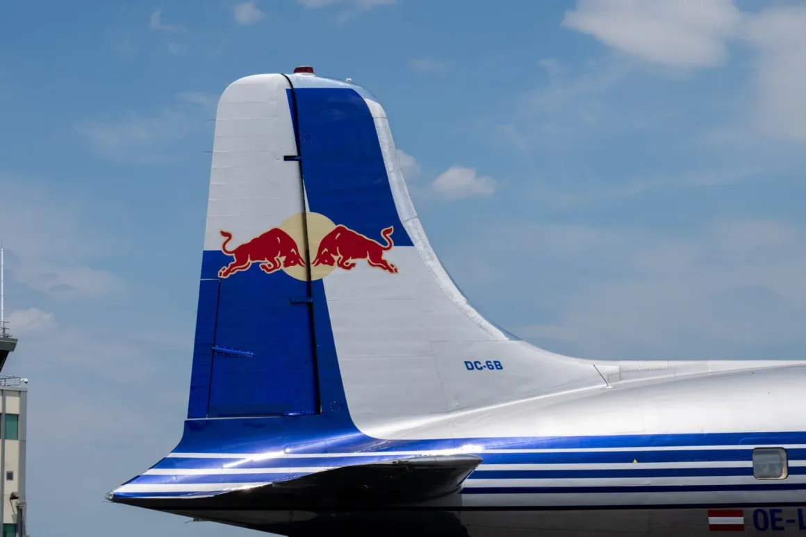 Red Bull, 68 Yıllık DC-6B'nin Tarihi Dönüşünü Vintage Bolex Kameralarla Belgeledi 2 Parçalı bulutlu gökyüzü önünde, kuyruğunda Red Bull logosu bulunan gümüş ve mavi renkli bir uçağın kuyruk bölümü. Uçağın üzerinde "DC-6B" ibaresi görülüyor.