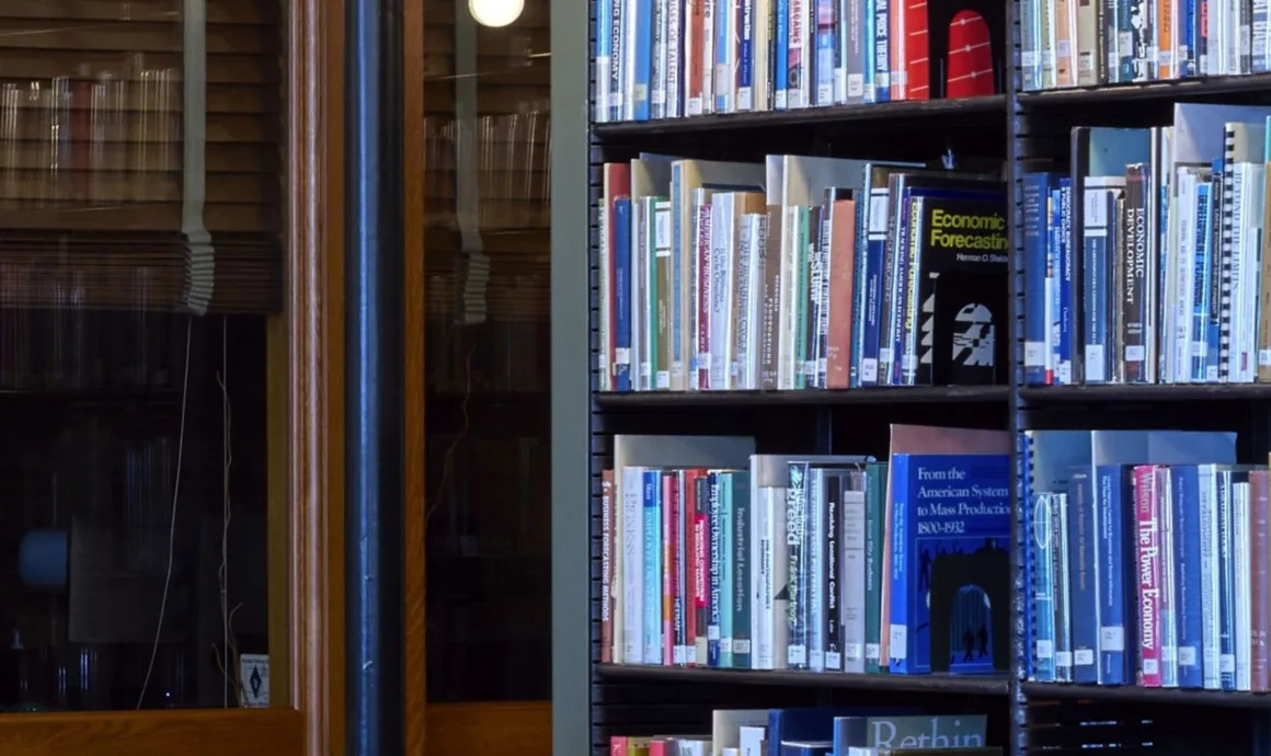 HiRezGo: Fotoğrafları Sıkıştırmadan Paylaşmanın Yeni Yolu 12 A bookshelf filled with various books stands next to a glass door with wooden frames, reflecting indoor lights. Some book titles are visible, and the setting suggests a quiet library or study area.