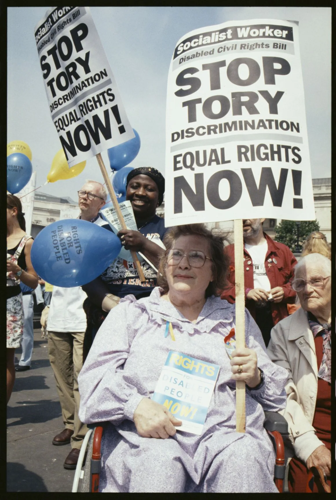 A woman in a wheelchair holds a placard reading "Stop Tory discrimination, equal rights now!" at a sunny Trafalgar Square rally, with balloons reading "Rights for disabled people" visible behind her.