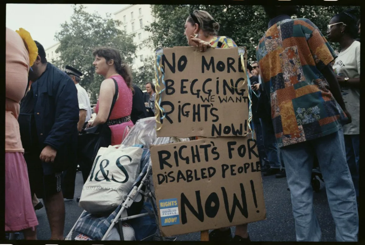 A protester holds two stacked cardboard signs reading "No more begging, we want rights now!" and "Rights for disabled people now!" at a busy central London street demonstration.
