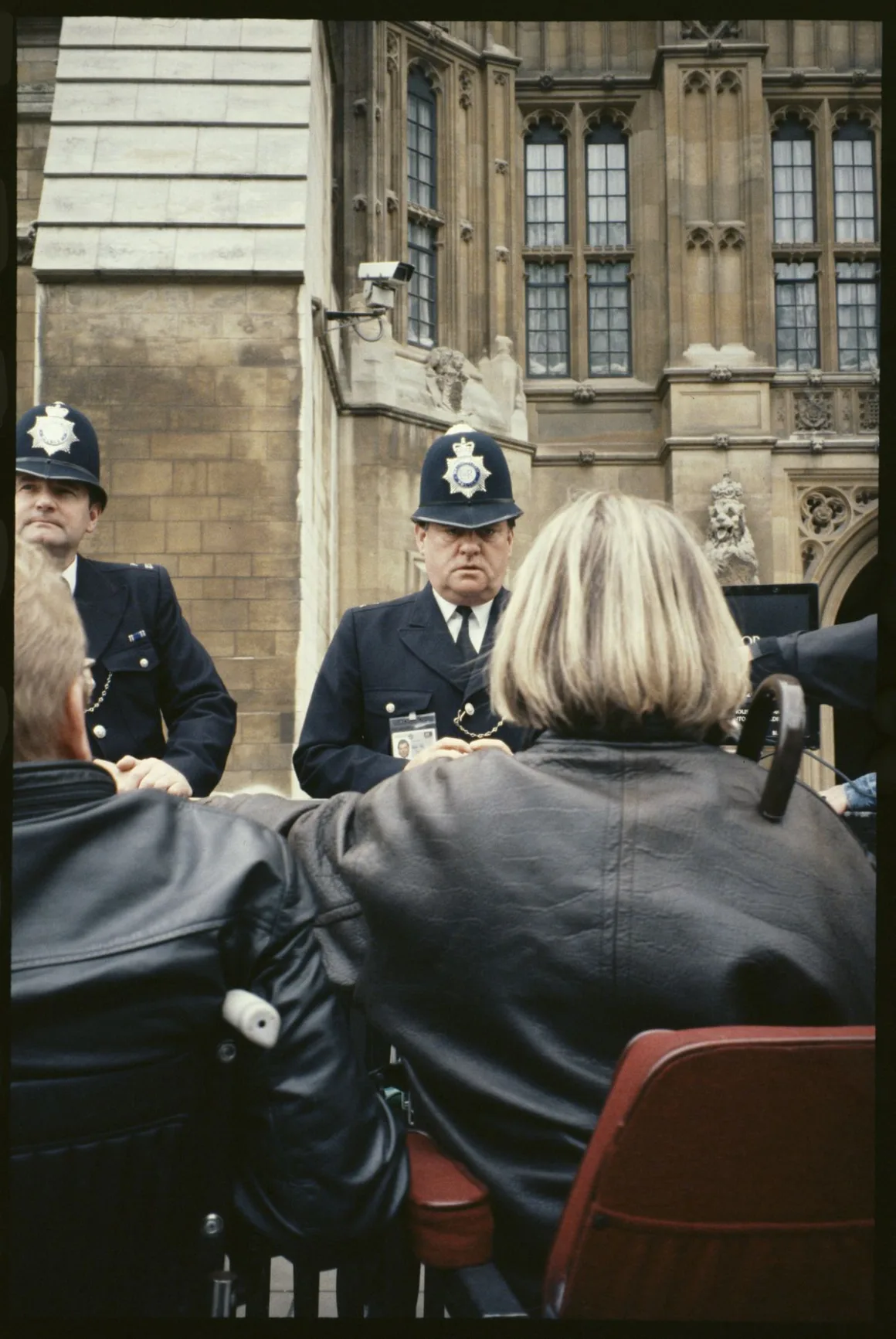 Two Metropolitan Police officers face a group of wheelchair users outside the Houses of Parliament, the Gothic stonework and a CCTV camera visible above them.