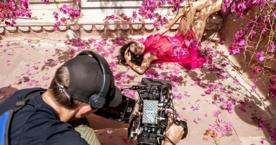 Canlı pembe bir elbise giyen bir kadın, pembe çiçek yapraklarıyla çevrili şekilde dekoratif taş bir duvarın önünde poz verirken bir fotoğrafçı çekim yapıyor.