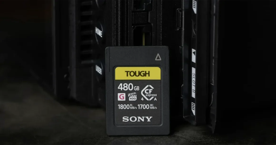 480 GB kapasiteli Sony TOUGH CFexpress Type A hafıza kartı, karanlık bir yüzey üzerinde elektronik bir cihazın (muhtemelen bir kamera veya kart okuyucunun) önünde dik şekilde duruyor.