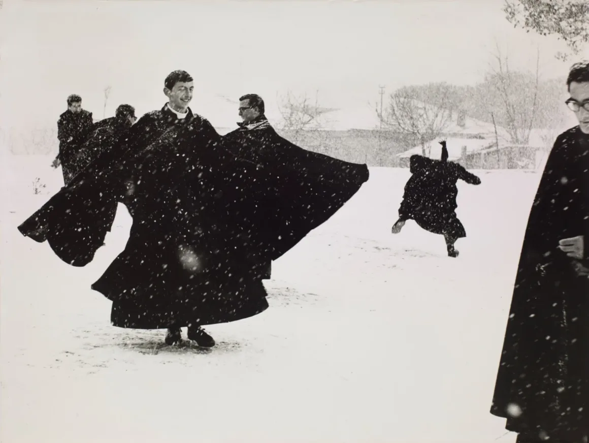 Several men in long black cassocks play in the snow, with one figure leaping in the background and another standing in the foreground.