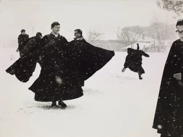 Several men in long black cassocks play in the snow, with one figure leaping in the background and another standing in the foreground.