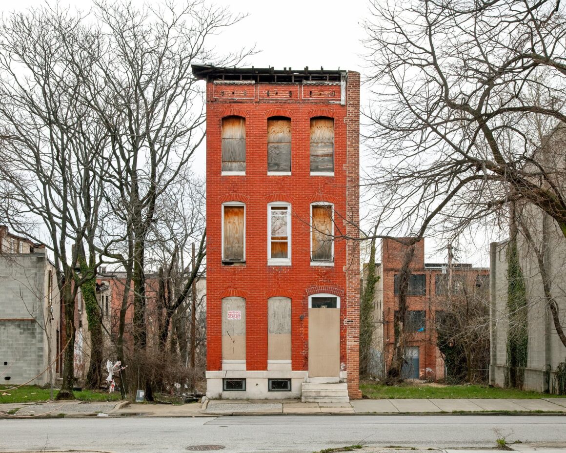 Ben Marcin, Baltimore’un Terk Edilmiş Evlerini 16 Yıldır Fotoğraflıyor 5 Dar ve üç katlı kırmızı tuğla bir bina, pencereleri ve kapıları tahtalarla kapatılmış halde, çıplak ağaçların arasında terk edilmiş olarak duruyor.