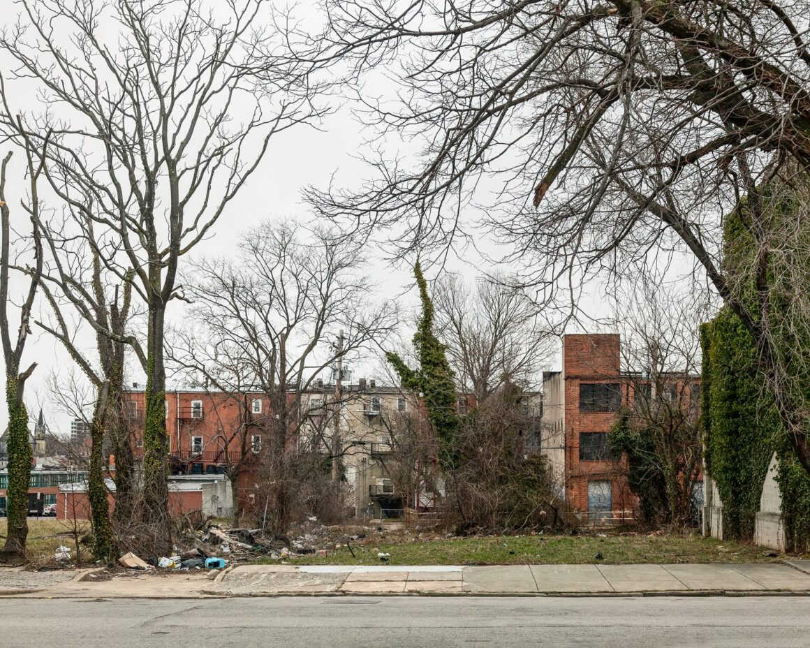 Ben Marcin, Baltimore’un Terk Edilmiş Evlerini 16 Yıldır Fotoğraflıyor 13 Tuğla apartmanların arasında, bulutlu bir gökyüzünün altında boş ve yabani otlarla kaplanmış bir arazi uzanıyor; ön planda çıplak ağaçlar ve etrafa saçılmış kalıntılar görülüyor.