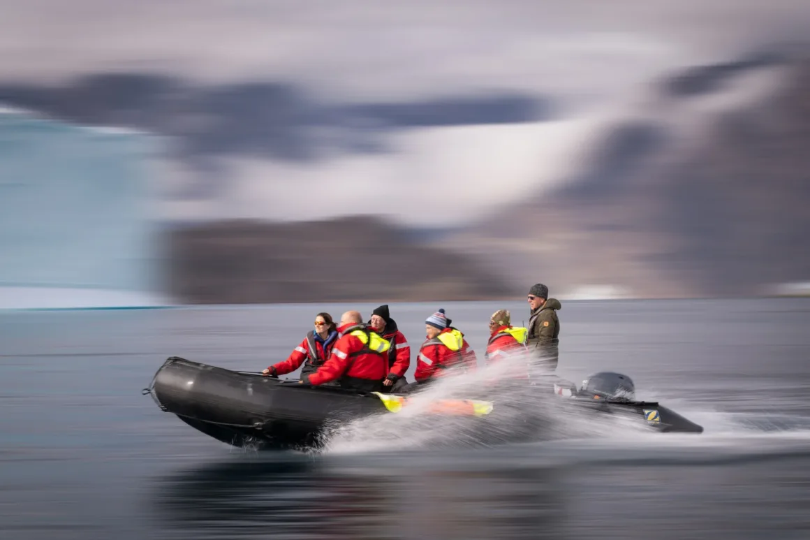 Scoresby Sund’da Işıklar Söndüğünde: Arktik’te Tam Güneş Tutulması 22 A group of people in red life jackets ride a black inflatable boat across the water with a blurred icy landscape behind.