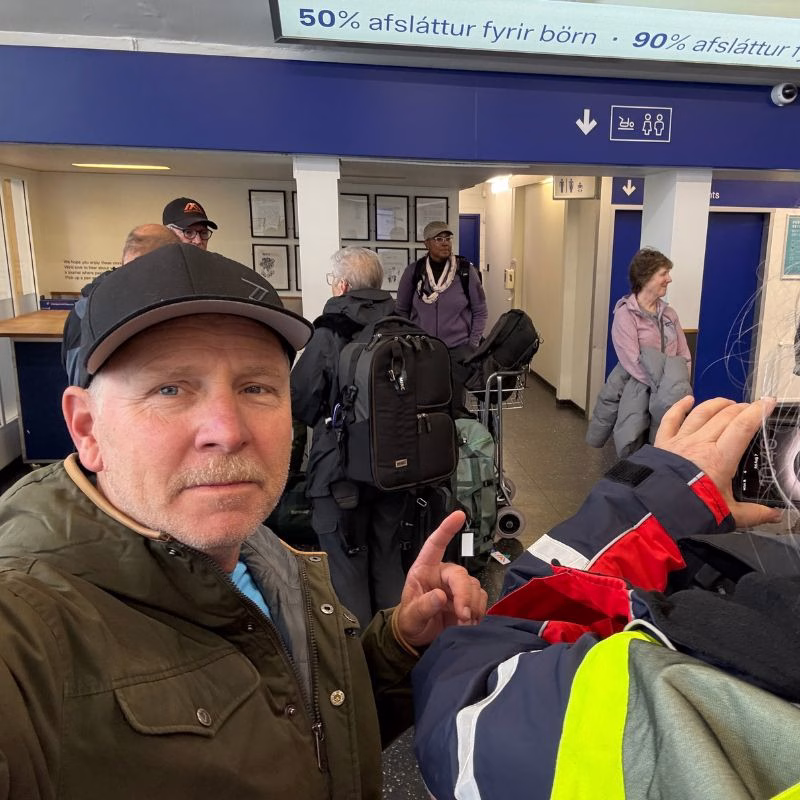 Scoresby Sund’da Işıklar Söndüğünde: Arktik’te Tam Güneş Tutulması 16 A man in a baseball cap takes a selfie in an airport terminal while another person in a high-visibility jacket points toward a restroom sign.