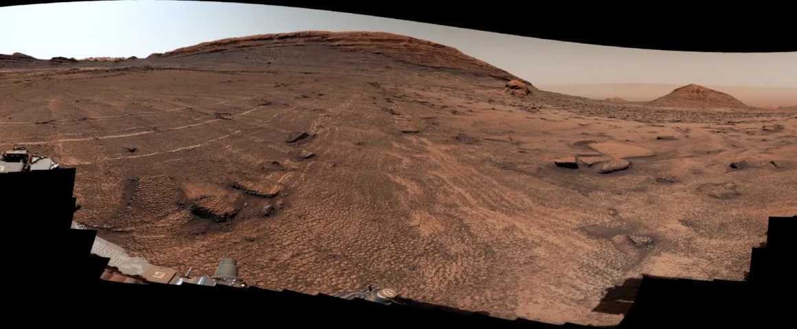 Ön planda keşif aracının ekipmanlarının, arka planda ise katmanlı büyük bir dağın yer aldığı kayalık ve çorak Mars arazisinin panoramik görünümü.