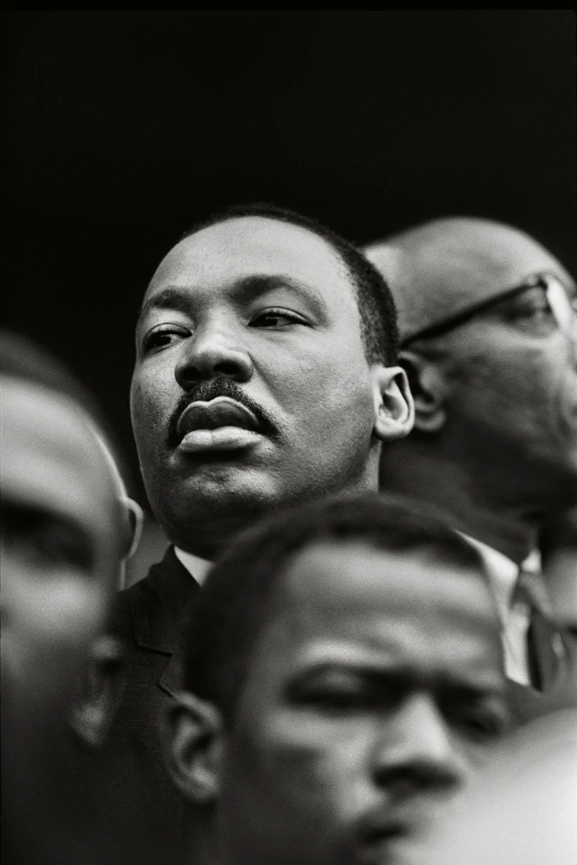Martin Luther King Jr.’ın kalabalıktaki diğer erkeklerle çevrili halde yana doğru baktığı yakın plan siyah-beyaz bir portre.