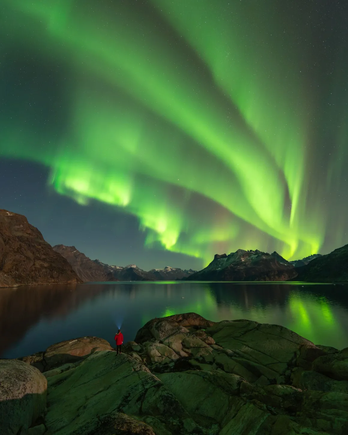 Dünyanın En Karanlık Gökyüzlerinin Güzelliğini Yansıtan 31 Ödüllü Fotoğraf 28 A person in a red jacket stands on rocky terrain at night, looking out over a calm lake toward mountains under a bright aurora.