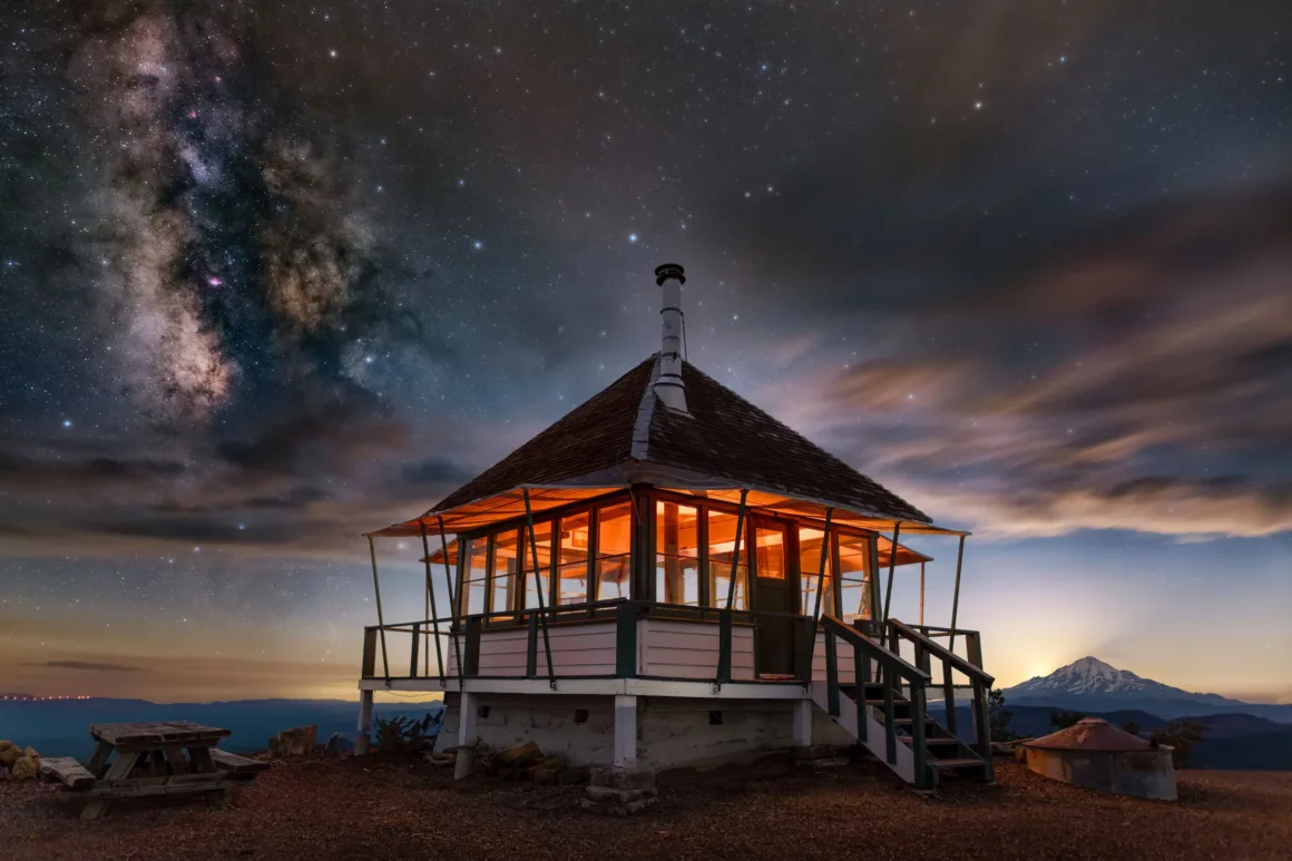 Dünyanın En Karanlık Gökyüzlerinin Güzelliğini Yansıtan 31 Ödüllü Fotoğraf 10 A wooden fire lookout cabin with glowing interior lights sits on a mountain peak under a starry night sky with the Milky Way.