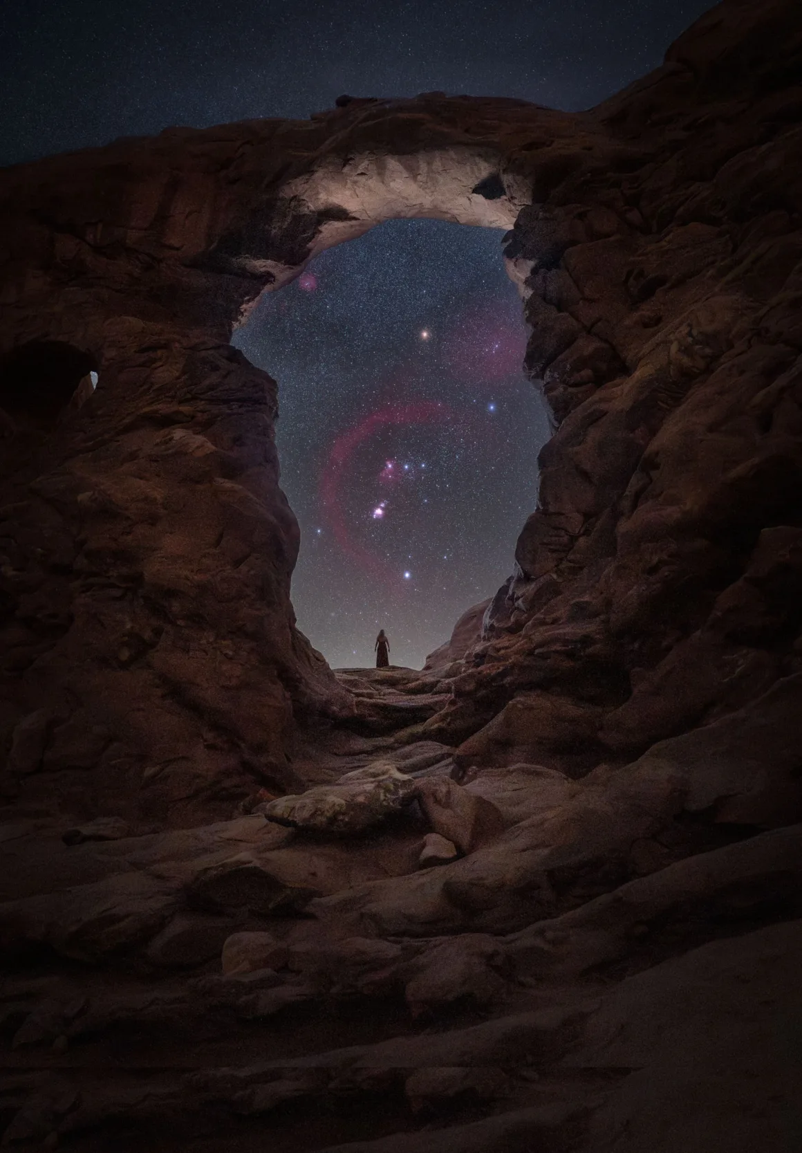 Dünyanın En Karanlık Gökyüzlerinin Güzelliğini Yansıtan 31 Ödüllü Fotoğraf 7 A person stands in the distance beneath a large natural rock arch, looking out at a night sky filled with stars and the Orion constellation.