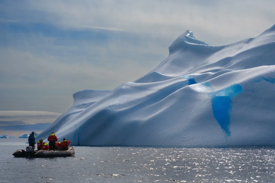 Scoresby Sund’da Işıklar Söndüğünde: Arktik’te Tam Güneş Tutulması 20 A small inflatable boat carrying several people in orange life vests floats near a massive, sculpted iceberg with bright blue ice patches.
