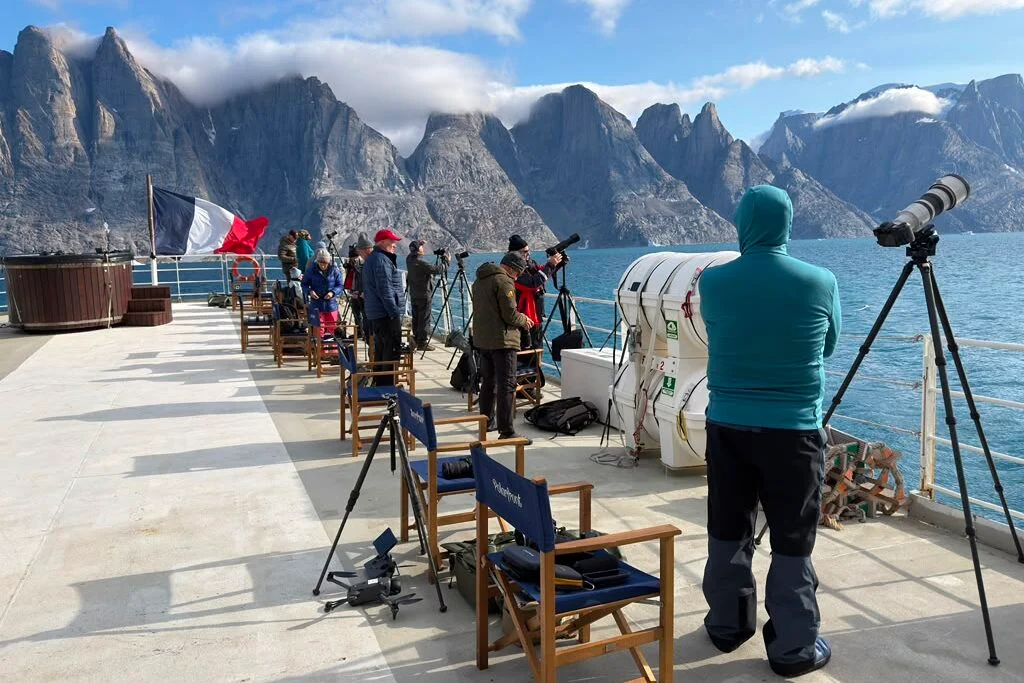 Scoresby Sund’da Işıklar Söndüğünde: Arktik’te Tam Güneş Tutulması 23 Bir grup insan, mavi gökyüzünün altındaki dağlık kıyıyı gözlemlemek için geminin güvertesinde tripodlara yerleştirilmiş fotoğraf makineleri ve gözlem dürbünleri kullanıyor.