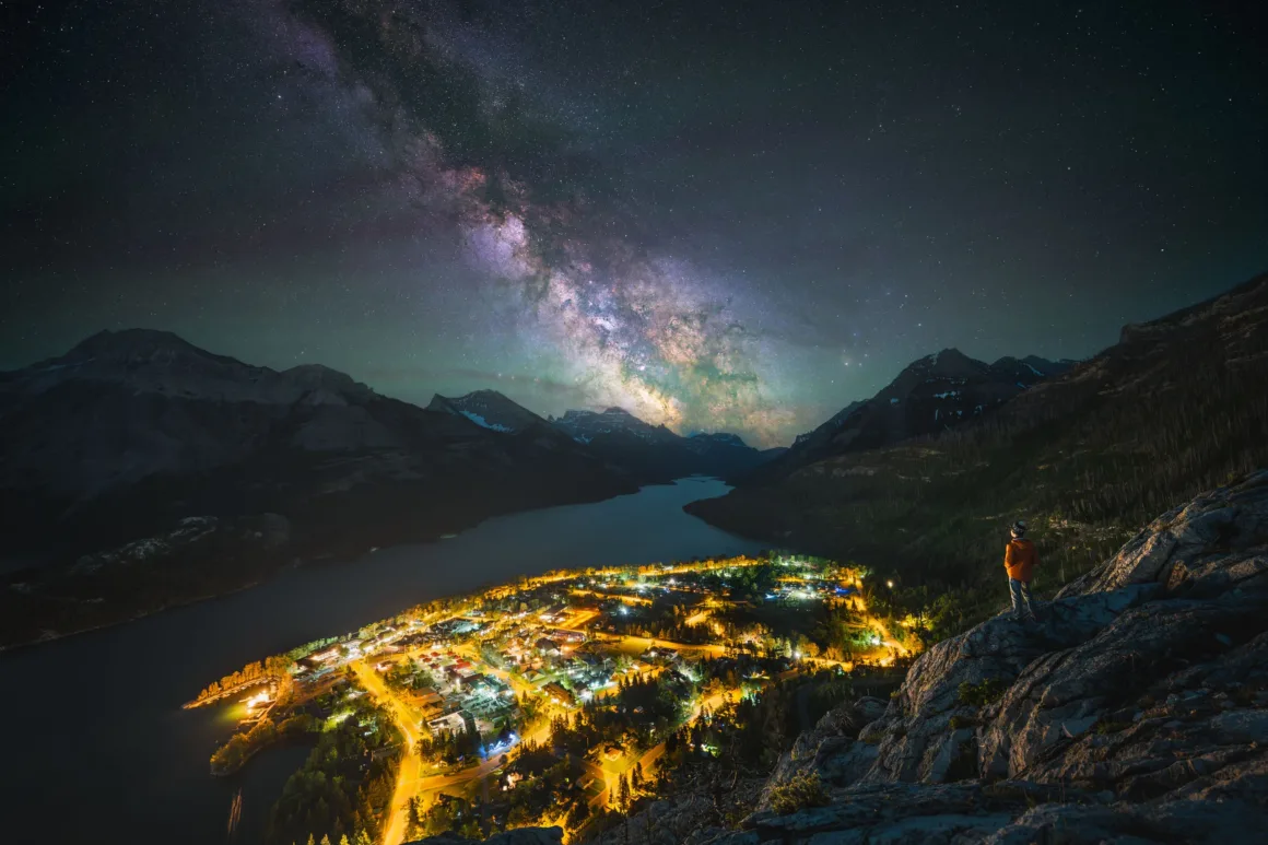 Dünyanın En Karanlık Gökyüzlerinin Güzelliğini Yansıtan 31 Ödüllü Fotoğraf 29 A person in an orange jacket stands on a rocky cliff overlooking a brightly lit town nestled beside a lake under a starry sky.