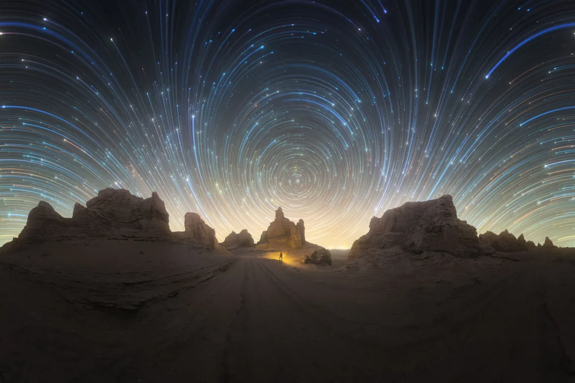 Dünyanın En Karanlık Gökyüzlerinin Güzelliğini Yansıtan 31 Ödüllü Fotoğraf 5 A wide-angle view of a desert landscape at night with a person standing near a rock formation under a sky filled with circular star trails.