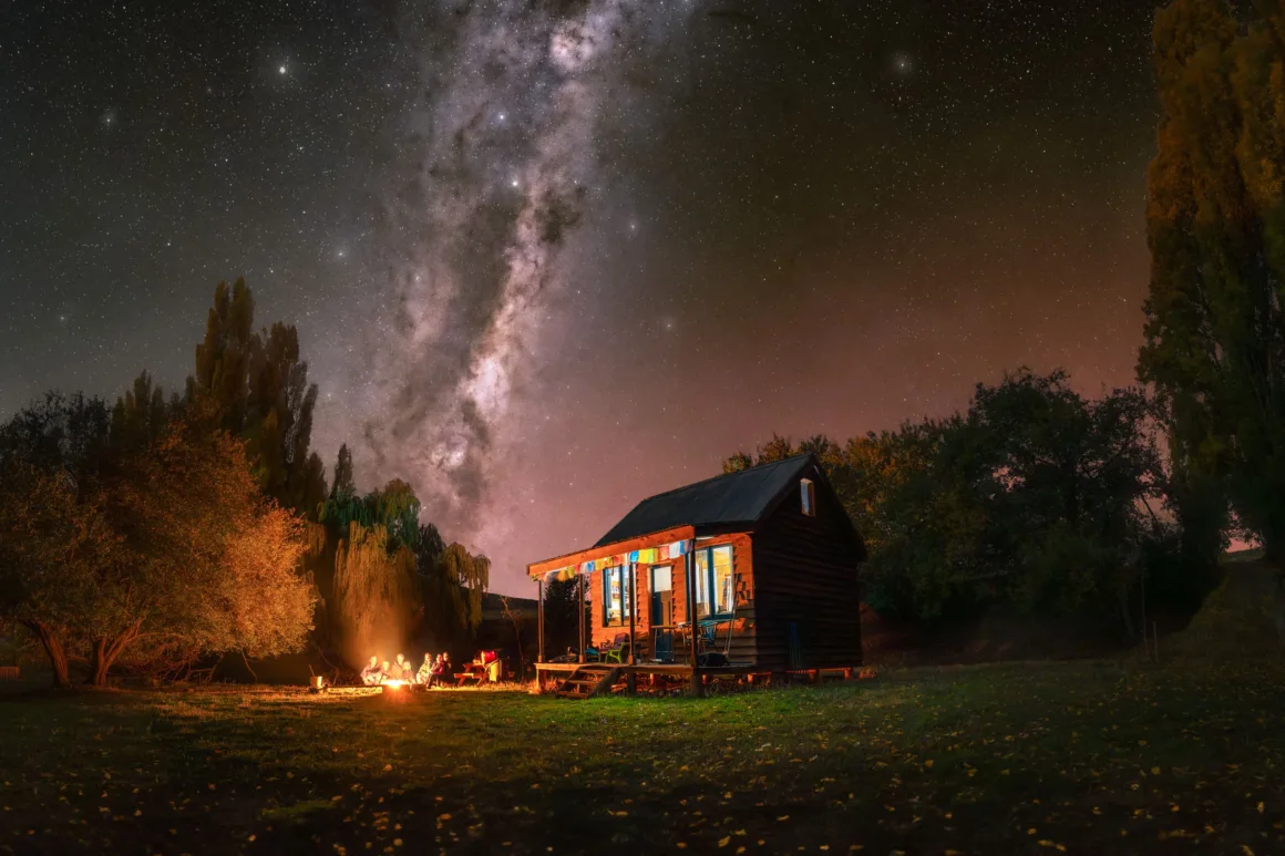 Dünyanın En Karanlık Gökyüzlerinin Güzelliğini Yansıtan 31 Ödüllü Fotoğraf 30 A small wooden cabin sits in a grassy field at night under a bright Milky Way, with a group of people gathered around a campfire.