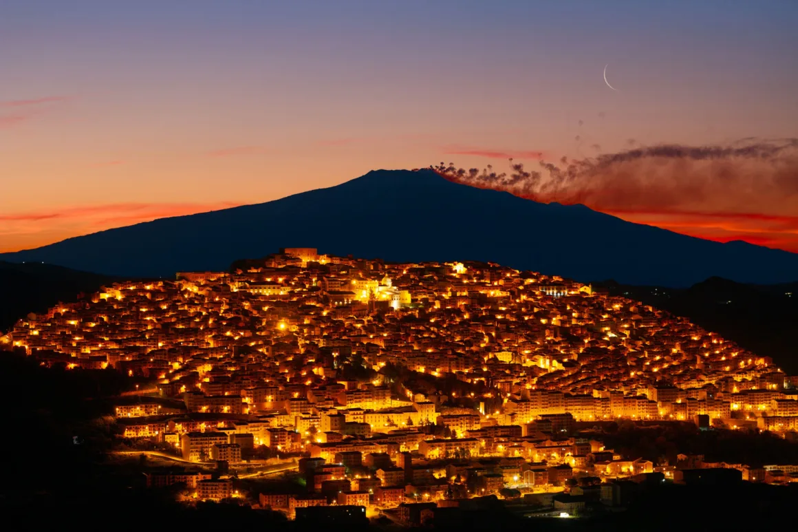 Dünyanın En Karanlık Gökyüzlerinin Güzelliğini Yansıtan 31 Ödüllü Fotoğraf 12 A densely packed town illuminated by warm lights at dusk, set against a large, smoking mountain and a thin crescent moon.
